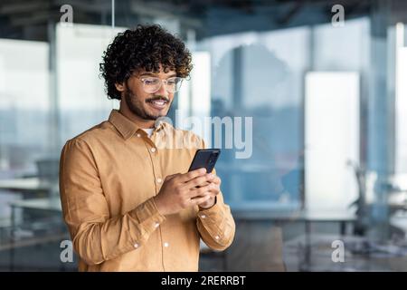 Hispanic man smiles and looks at the phone screen, businessman inside the office holds a smartphone, browses online internet pages, makes a call, and uses an application, near the window in a shirt. Stock Photo