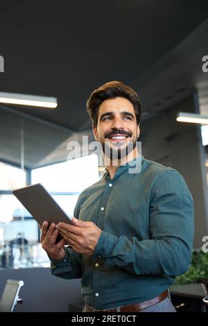 Happy young Latin business man holding tablet standing in office looking away. Stock Photo
