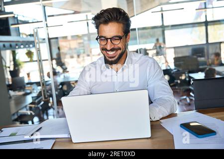 Happy smiling young Latin business man working in office looking at laptop. Stock Photo
