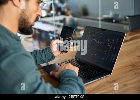Investor holding phone using laptop analyzing stock trade market data. Stock Photo