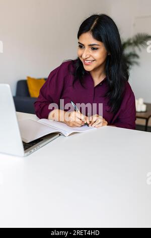 Young adult indian student woman taking notes while using laptop at home Stock Photo