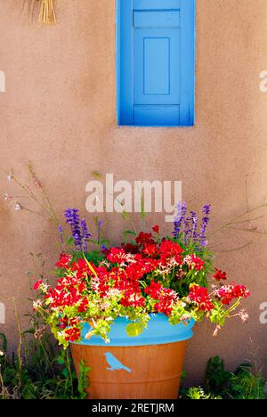 Ranchos De Taos, New Mexico, USA - July 23, 2023: Potted flowers bloom under a blue painted wood shutter on the wall of a traditional adobe home. Stock Photo