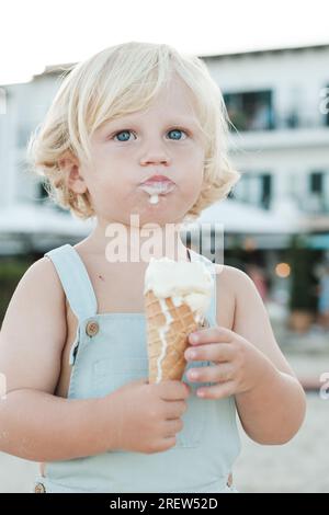 Fair haired child eating ice cream cone while standing on blurred street with smeared mouth in daylight Stock Photo