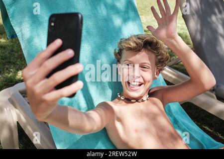 Happy boy with blond hair sitting on blue blanket and looking at screen of mobile phone while laughing with opened mouth and saying hello with raised Stock Photo