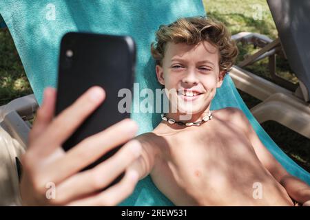 Cheerful blond hair boy smiling and looking at screen of mobile phone while taking selfie in blanket chair in resort garden Stock Photo