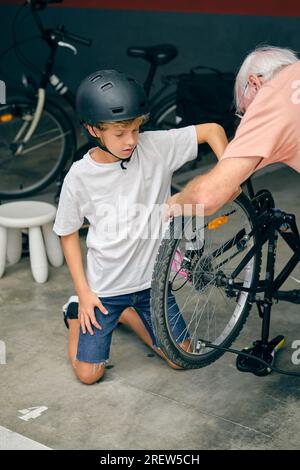 High angle of curious boy in protective helmet sitting on knees and fixing bicycle with old grandfather in garage Stock Photo