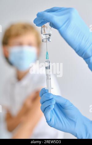 Crop unrecognizable medic in sterile gloves with small bottle filling injector with COVID 19 vaccine against patient in clinic Stock Photo