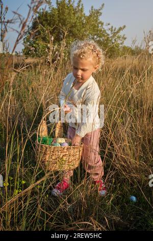 Funny little girl in white sweater collecting dyed Easter eggs into wicker basket on grassy verdant meadow in sunny spring nature Stock Photo