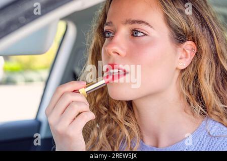 Crop sensitive female driver with blue eyes and wavy hair applying red lipstick in car on sunny day Stock Photo