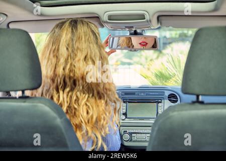 Back view young female driver applying red lipstick against vanity mirror on sun visor whiles sitting in car drivers seat on sunny weather Stock Photo