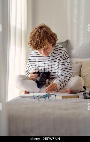 Content schoolchild with professional photo camera sitting with crossed legs near notepad on bed in house Stock Photo