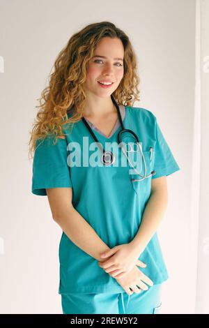 Positive young female physician with long wavy hair in uniform and stethoscope standing near white wall with crossed hands and looking at camera Stock Photo