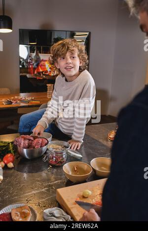 Curly smiling boy in sweater sitting on wide kitchen counter full of kitchenware against father Stock Photo
