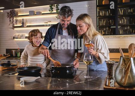 Positive parents in aprons preparing traditional dish with curly haired son in kitchen and drinking wine while spending time together Stock Photo