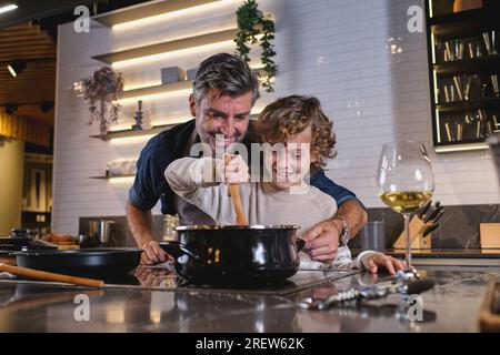 Happy curly haired boy stirring hot dish in saucepan and helping middle aged father with cooking while spending time together in kitchen at home Stock Photo