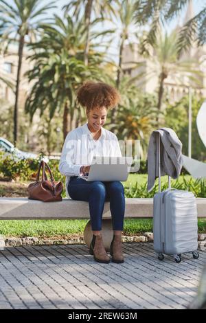 Full body of content African American lady sitting on bench in sunny park and working on remote project on laptop Stock Photo