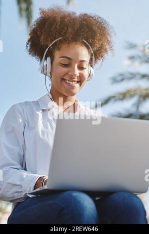 Low angle of cheerful African American female freelancer in headset smiling while working remotely on netbook and listening to music in park on blurre Stock Photo