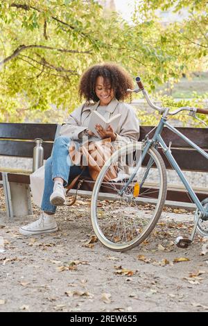 Full body of young smiling ethnic female student with curly hair sitting on bench in park near bike and reading book while doing homework Stock Photo