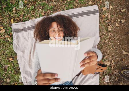 Top view of black woman with Afro hairstyle reading interesting story and smiling while resting on blanket on grass in nature Stock Photo