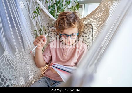Concentrated child in casual clothes and glasses writing in notepad while resting in knitted hammock and doing homework on balcony at home Stock Photo
