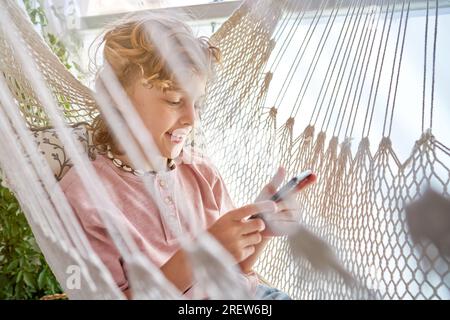 Side view of smiling curly haired boy relaxing in wicker hammock and browsing internet on cellphone on balcony with plants Stock Photo