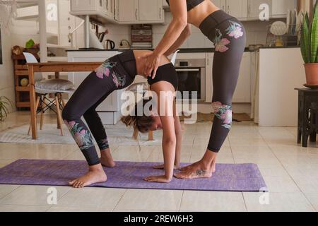Side view of crop mother helping daughter to stand in Urdhva Dhanurasana while practicing yoga together on mat in modern apartment Stock Photo
