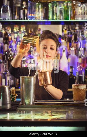 Confident young female bartender with dark hair sprinkling salt into cup while mixing flaming cocktail at counter in modern bar Stock Photo