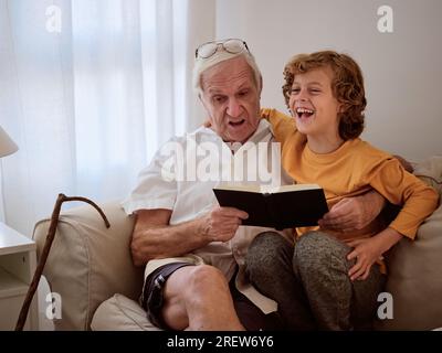 Senior man and happy grandson sitting on sofa and reading interesting book while spending time together at home Stock Photo