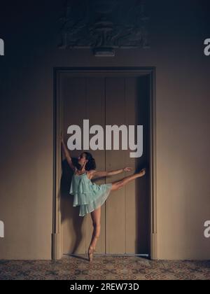 Side view of flexible lady in summer dress and pointe shoes practicing arabesque against room door in dim light Stock Photo