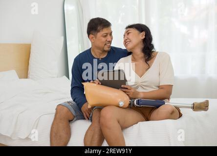 A man and a woman engrossed in a digital tablet while sitting on bed. A prosthetic bionic leg rests on her  lap. Stock Photo