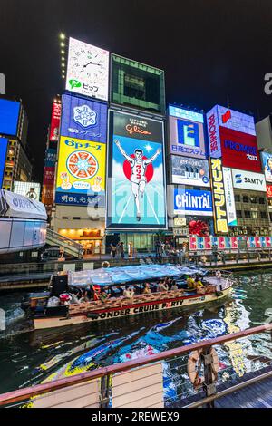 View from the Ebisu bridge at night of the famous Glico Running Man neon light display at Dotonbori, Osaka, with a tourist cruise boat passing. Stock Photo