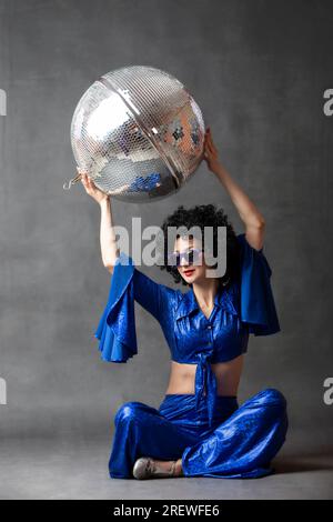 A woman in a seventies flared suit with a disco ball in an afro wig on a gray background. Stock Photo