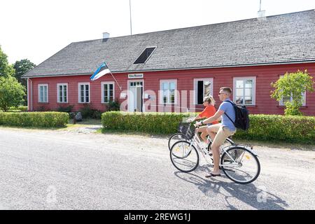 Tourists on bicycles cycling passing beautiful traditional wooden building of a kihnu museum on remote Kihnu island in baltic sea, Estonia, Stock Photo