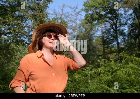 A woman in a shady green park. Orange dress, straw hat, sunglasses. Place for text Stock Photo