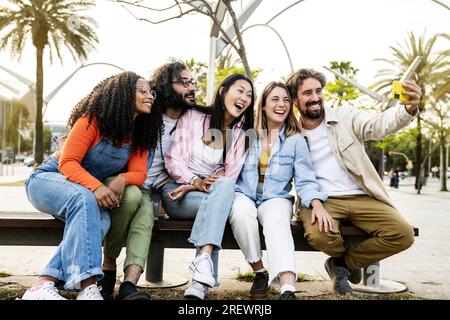 Diverse group of friends taking a selfie in the street sitting on a bench. Cheerful multiracial group of young hipsters taking a picture outside in a Stock Photo