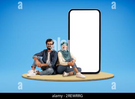 Cheerful muslim couple sitting by big smartphone with white screen Stock Photo