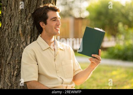 Closeup young man sitting under tree at park, reading book Stock Photo
