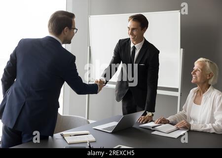 Two positive successful businessmen shaking hands, standing at table Stock Photo