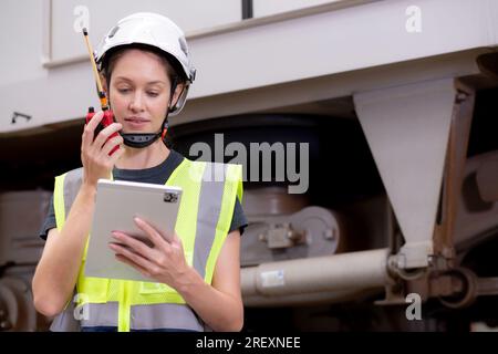 Young caucasian engineer woman using radio for command with worker checking electric train for planning maintenance and looking digital tablet in stat Stock Photo