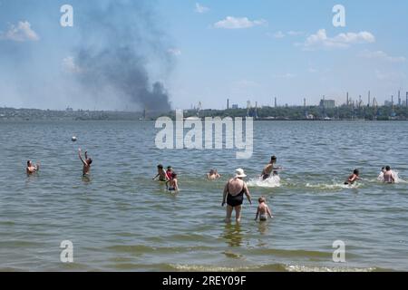 People bathing in the Dnieper River as industrial plants were hit by a rocket in the background. Despite the currently ongoing Ukrainian counteroffensive in the Zaporizhzhya region, and the risk of a terrorist attack on the nuclear power plant, daily life in the Zaporizhzhia region is still going on. Warm evenings fill the city's streets with strolling people, while sunflowers are blooming in the frontline areas and the first harvests has begun. However, intensified warfare does not let up - intense shelling almost daily takes the lives of people, who additionally suffer from lack of water aft Stock Photo