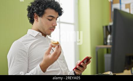 Young latin man business worker having rest eating muffin using smartphone at office Stock Photo