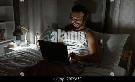 Young arab man using laptop sitting on the bed wearing headphones at bedroom Stock Photo