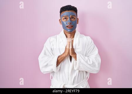 Young hispanic man wearing beauty face mask and bath robe begging and praying with hands together with hope expression on face very emotional and worr Stock Photo