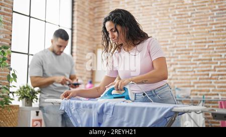 Man and woman couple ironing clothes while boyfriend using smartphone at laundry room Stock Photo
