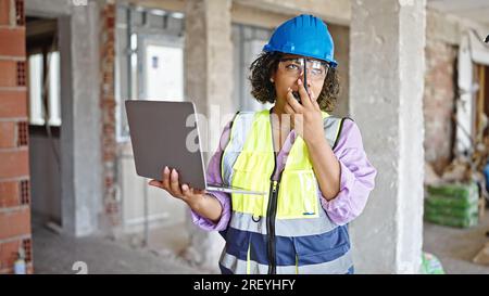 Young beautiful latin woman builder using laptop talking by walkie talkie at construction site Stock Photo