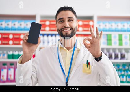 Hispanic young man working at pharmacy drugstore showing smartphone screen doing ok sign with fingers, smiling friendly gesturing excellent symbol Stock Photo