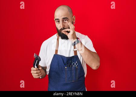 Young hispanic man with beard and tattoos wearing barber apron holding razor mouth and lips shut as zip with fingers. secret and silent, taboo talking Stock Photo