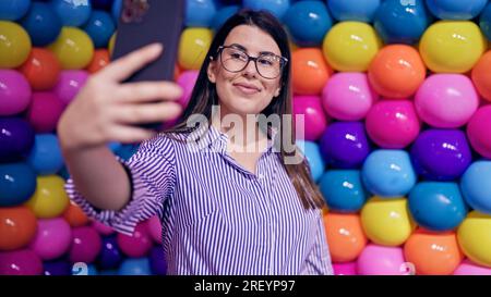 Young beautiful hispanic woman visiting colorful futuristic exhibition space taking selfie Stock Photo