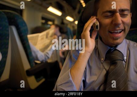 A man talking on the phone in a train compartment, with a passenger reading a newspaper in the background. Stock Photo