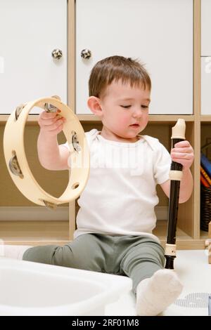 Toddler baby plays the flute sitting on the floor in the children's room. Child boy playing musical instruments. Kid aged one year eight months Stock Photo